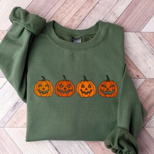 May include: A green sweatshirt with four orange jack-o'-lanterns with different carved faces. The pumpkins are outlined in black and have varying expressions, ideal for Halloween. The sweatshirt is made of a soft material.