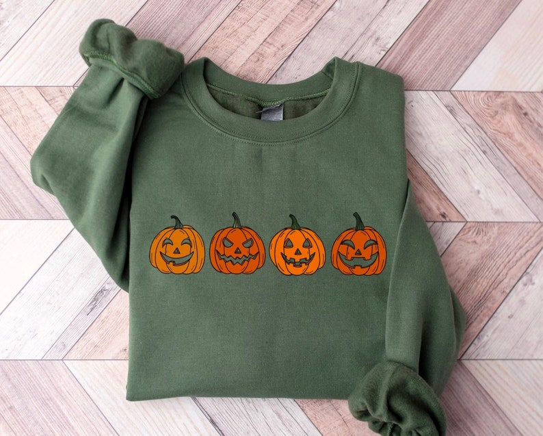 May include: A green sweatshirt with four orange jack-o'-lanterns with different carved faces. The pumpkins are outlined in black and have varying expressions, ideal for Halloween. The sweatshirt is made of a soft material.