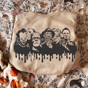 May include: Beige crewneck sweatshirt featuring a black graphic of iconic horror movie characters, including Pennywise, Jason Voorhees, Frankenstein's monster, Freddy Krueger, and Michael Myers. The design has a dripping effect at the bottom. The words "Class Design Company" are printed on the sweatshirt.