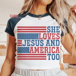 May include: White t-shirt with a blue and red American flag design. The text "She Loves Jesus and America Too" is printed on the shirt.