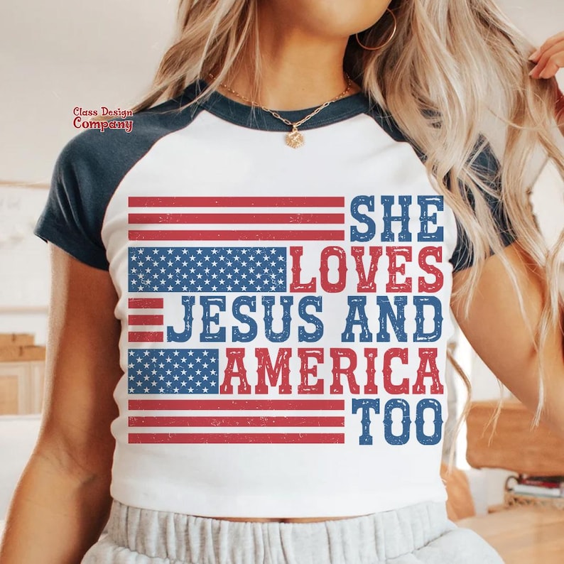 May include: White t-shirt with a blue and red American flag design. The text "She Loves Jesus and America Too" is printed on the shirt.