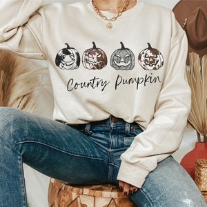 May include: A cream-coloured sweatshirt displaying four pumpkin illustrations with different patterns and expressions. The words "Country Pumpkin" are written underneath in a cursive style. The person is wearing blue jeans.