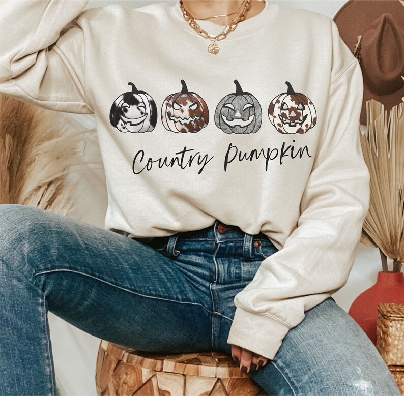 May include: A cream-coloured sweatshirt displaying four pumpkin illustrations with different patterns and expressions. The words "Country Pumpkin" are written underneath in a cursive style. The person is wearing blue jeans.