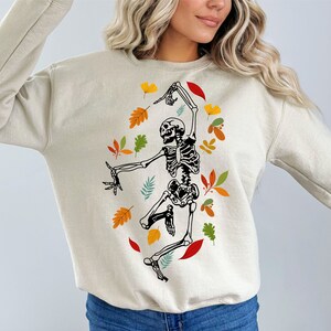 May include: Cream-coloured sweatshirt with a dancing skeleton design, encircled by autumnal leaves. The skeleton is black and white, with detailed bone structure. The leaves are in shades of red, orange, yellow, and green, creating a seasonal theme.