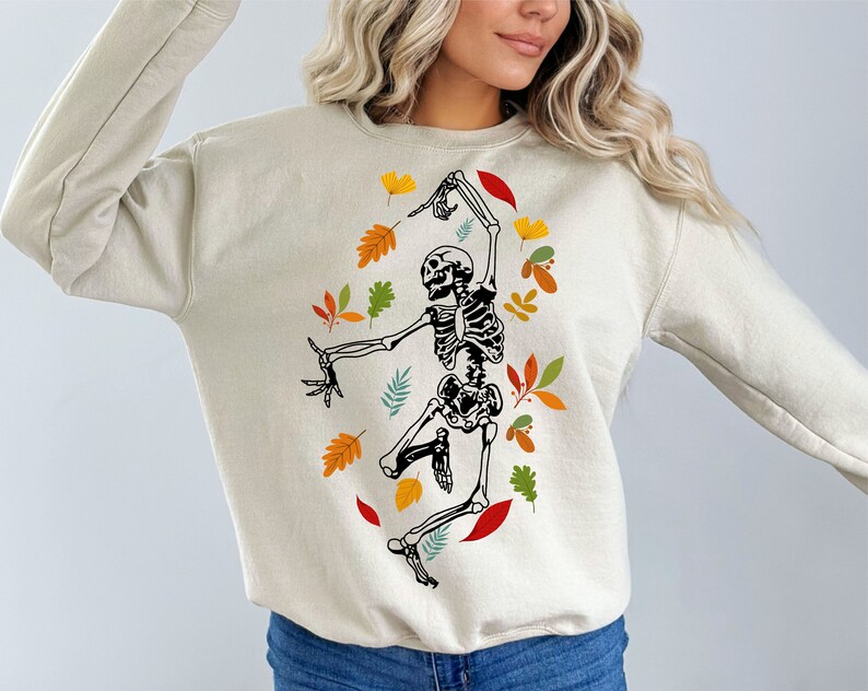 May include: Cream-coloured sweatshirt with a dancing skeleton design, encircled by autumnal leaves. The skeleton is black and white, with detailed bone structure. The leaves are in shades of red, orange, yellow, and green, creating a seasonal theme.