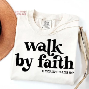 May include: A white t-shirt with black text that reads "walk by faith 2 Corinthians 5:7".