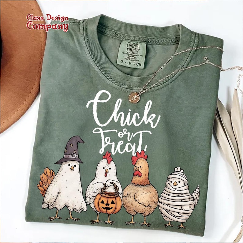 May include: Green t-shirt with the words "Chick or Treat" in white script. The shirt features four cartoon chickens dressed in Halloween costumes, including a witch, a pumpkin carrier, a rooster, and a mummy. A silver necklace is draped across the shirt.