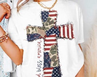 Puede incluir: Una camiseta blanca de manga corta con un gráfico de cruz grande hecho de patrones de tela de retazos, incluyendo rayas rojas y blancas, azul con estrellas blancas y camuflaje verde. El texto god bless america está impreso en rojo.