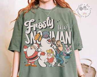 May include: A sage green short-sleeve t-shirt featuring a graphic print of Frosty the Snowman, Santa Claus, and other characters. The text reads Frosty the Snowman. The design includes stars and a circular logo in the top right corner.