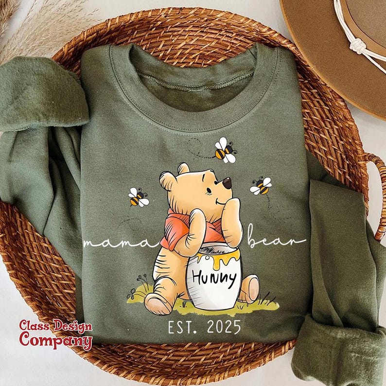 May include: Green sweatshirt with a cartoon Winnie the Pooh holding a jar of honey that says "Hunny". The text "mama bear" and "EST. 2025" are printed on the sweatshirt. There are bees flying around Winnie the Pooh.