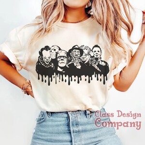 May include: Cream-colored t-shirt featuring a black graphic of iconic horror movie characters, including Pennywise, Jason Voorhees, and Freddy Krueger. The design has a dripping effect at the bottom. The text "Class Design Company" is printed on the bottom right.