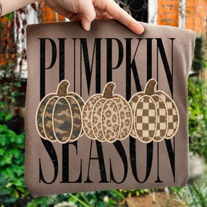 May include: A brown t-shirt featuring the black text PUMPKIN SEASON and three pumpkin illustrations. The first pumpkin has a camouflage pattern, the second has a leopard print, and the third has a brown and tan checkered pattern.