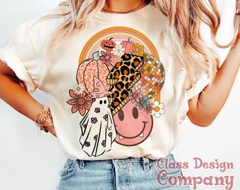Può includere: Una t-shirt color crema con una stampa grafica raffigurante un fantasma con cappello da strega, un fulmine leopardato, una faccina sorridente rosa, zucche e una palla da discoteca sotto un arcobaleno multicolore con fiori.