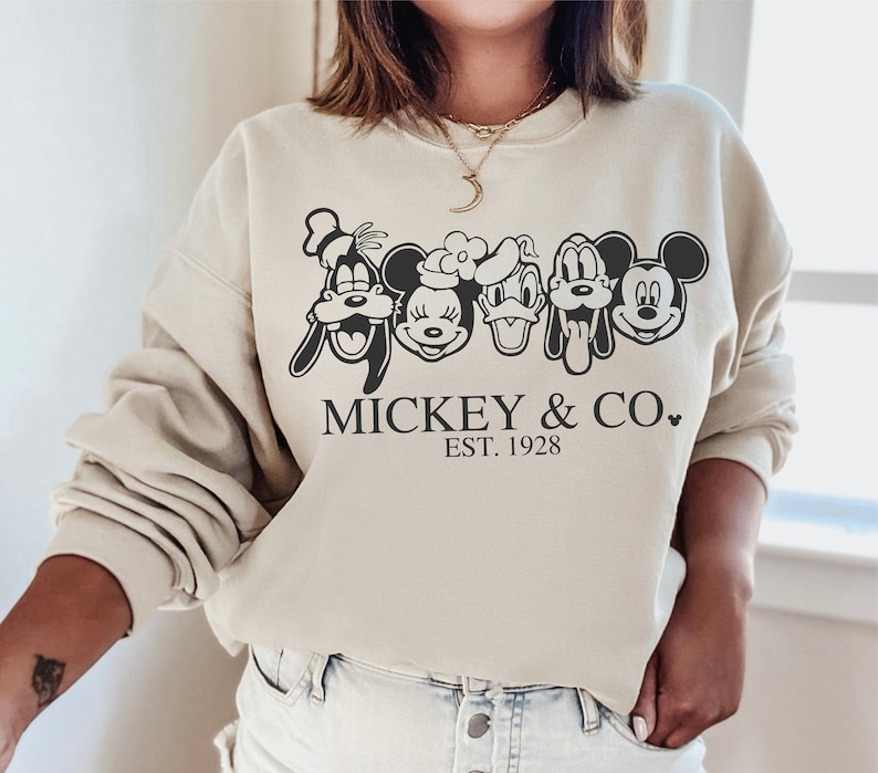 K&ouml;nnte beinhalten: Ein beiger Pullover mit einer schwarzen Grafik von Mickey Mouse, Minnie Mouse, Donald Duck, Goofy und Pluto. Der Text "Mickey & Co. EST. 1928" ist unter den Figuren gedruckt.