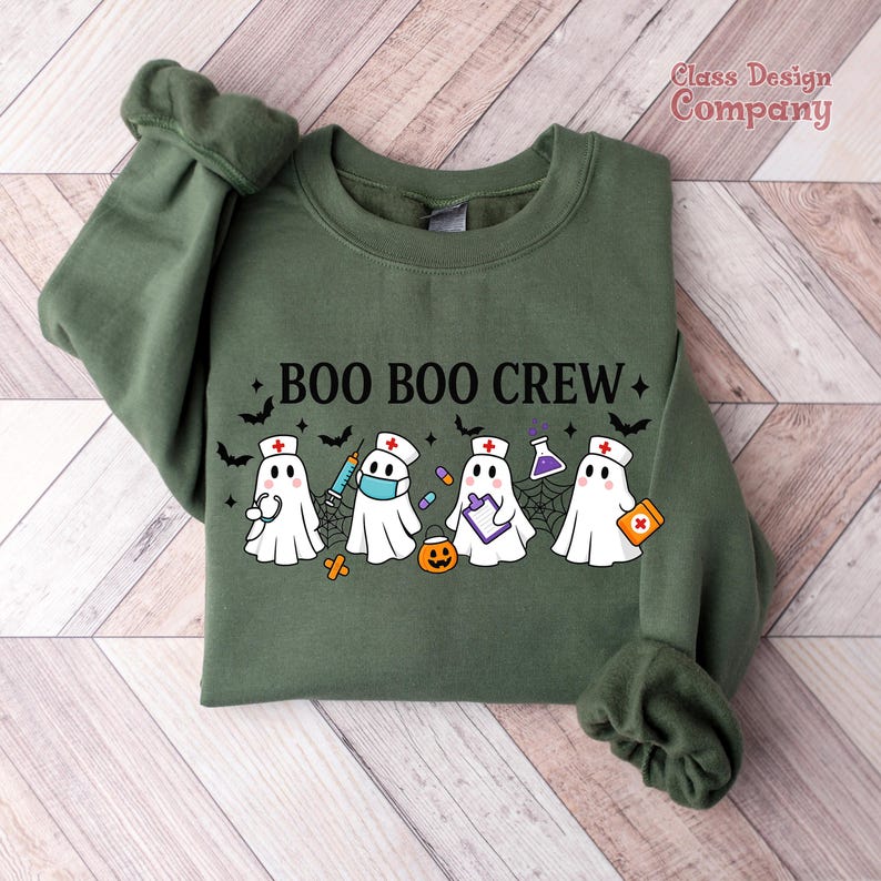 May include: An olive green sweatshirt with the text "BOO BOO CREW" and illustrations of ghosts dressed as medical professionals. The ghosts are wearing nurse hats, face masks, and holding medical supplies. Bats and stars are also featured.