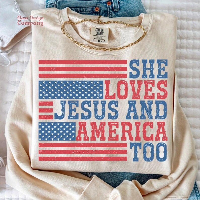 May include: A cream-coloured sweatshirt with a red, white and blue American flag design. The text "SHE LOVES JESUS AND AMERICA TOO" is printed in blue and red on the sweatshirt.