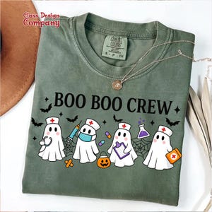May include: Green t-shirt with the text "BOO BOO CREW" and illustrations of ghost characters dressed as nurses, with bats, spiderwebs, and a jack-o'-lantern. The shirt has a Comfort Colors label.