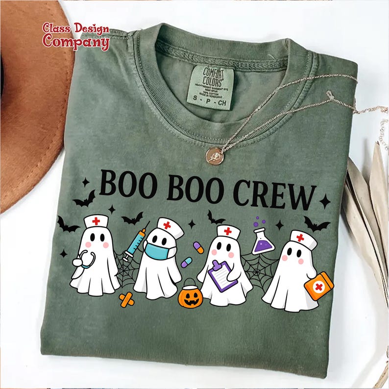 May include: Green t-shirt with the text "BOO BOO CREW" and illustrations of ghost characters dressed as nurses, with bats, spiderwebs, and a jack-o'-lantern. The shirt has a Comfort Colors label.
