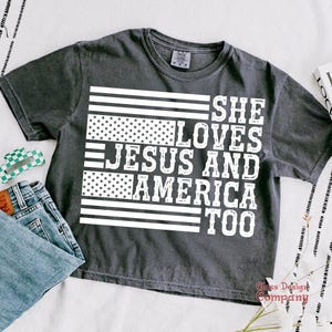 May include: A dark grey t-shirt with a white graphic design of an American flag and the text "She Loves Jesus and America Too".