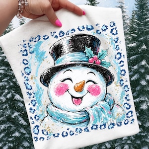 May include: A white t-shirt featuring a graphic illustration of a smiling snowman wearing a black top hat with holly berries, a light blue scarf, and pink cheeks, all framed by a blue leopard print border with gold paint splatters.