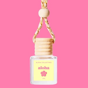 May include: A clear glass bottle with a wooden cap and a yellow label that says "Aloha Collectives aloha" with a pink flower graphic. The bottle is hanging from a string with a wooden bead.