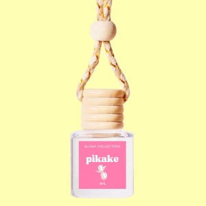 May include: A small glass bottle with a wooden cap and a pink label that says "Aloha Collectives pikake 8ML". The bottle is hanging from a beige and gold rope.