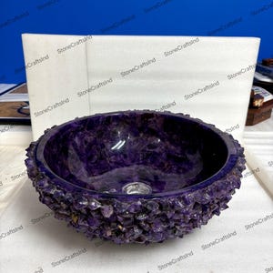 May include: A round, purple amethyst stone sink with a rough, textured exterior. The interior is smooth and polished, showcasing the natural color variations of the gemstone. A drain hole is visible in the center.