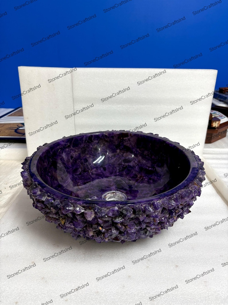 May include: A round, purple amethyst stone sink with a rough, textured exterior. The interior is smooth and polished, showcasing the natural color variations of the gemstone. A drain hole is visible in the center.