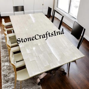 May include: A rectangular dining table with a white, textured surface, surrounded by chairs. The table has a decorative base and is in a well-lit room. The text "StoneCraftsInd" is visible on the table.
