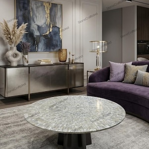 May include: A circular coffee table with a mother-of-pearl surface and a dark brown base. The table is in a modern living room with a purple sofa, a silver cabinet, and an abstract painting.