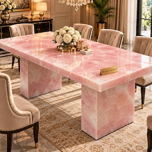 Può includere: Un tavolo da pranzo rettangolare in quarzo rosa lucido con una superficie liscia e riflettente, sostenuto da due spesse basi a piedistallo in pietra abbinata. Una piccola targhetta dorata sul tavolo riporta il testo StoneCraftsInd.