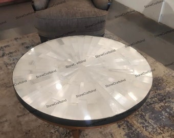 May include: A round white coffee table with a unique design made of white stone pieces. The table has a black rim and sits on four wooden legs.