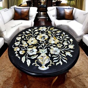 May include: A round black coffee table with a floral inlay design. The table features a dark wood base and a detailed inlay of flowers and leaves in shades of white, cream, and gold. The table is a decorative piece for a living room.
