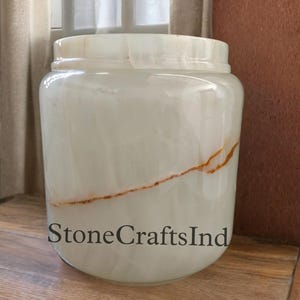 Könnte beinhalten: Weiße Onyx-Vase mit braunem Streifen. Die Vase ist rund und hat eine glatte, polierte Oberfläche. StoneCraftsInd ist auf die Vase gedruckt.