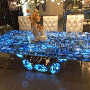 Puede incluir: Una mesa de comedor de ágata azul con un acabado de epoxi transparente pulido. La superficie de la mesa está hecha de rebanadas de ágata natural, creando un patrón único y hermoso. La mesa está soportada por una base de metal con un diseño geométrico. La mesa está iluminada desde abajo, creando un efecto impresionante.
