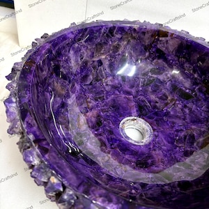 May include: A polished, oval-shaped amethyst sink. The sink's interior is a deep purple with swirling patterns, and the rim is encrusted with raw amethyst crystals. A drain hole is visible at the bottom. The sink is a unique bathroom fixture.