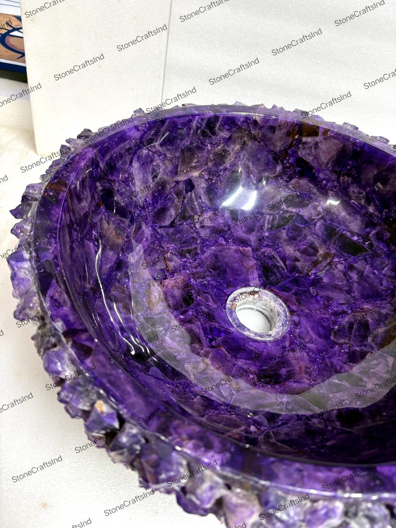 May include: A polished, oval-shaped amethyst sink. The sink's interior is a deep purple with swirling patterns, and the rim is encrusted with raw amethyst crystals. A drain hole is visible at the bottom. The sink is a unique bathroom fixture.