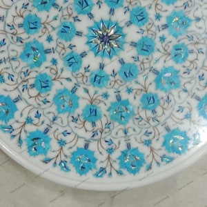 May include: A white marble table top with a floral design. The design features turquoise blue flowers and leaves with silver accents. The table has a white border and a polished finish. The overall theme is decorative and elegant.