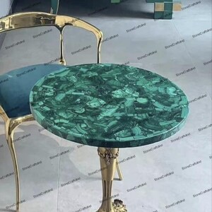 Op de afbeelding: Een ronde groene malachiet tafelblad met gouden metalen poten. Het tafelblad heeft een uniek patroon van groene en zwarte swirls.