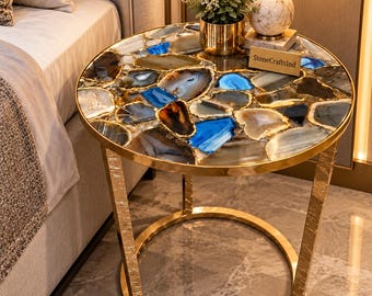 May include: A round side table featuring a tabletop made of multicolored agate slices in shades of blue, gray, and brown with a gold-toned metal frame. The table has a circular base and textured legs. A small rectangular sign reads StoneCraftsInd.