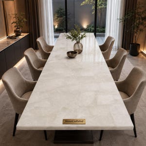Op de afbeelding: Een lange rechthoekige eettafel met een wit stenen oppervlak met een geometrisch tegelpatroon en een koperen plaatje met de tekst StoneCraftsInd. De tafel is omringd door zes beige gestoffeerde fauteuils met donkerbruine taps toelopende poten.