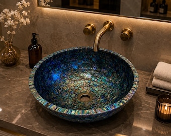 May include: A round vessel sink featuring a mosaic of iridescent blue and green abalone shell fragments. The sink is paired with a wall-mounted brushed gold faucet and two matching circular control knobs. A small gold plaque reads StoneCraftsInd.