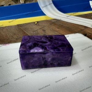 Puede incluir: Una caja rectangular de piedra púrpura con tapa. La caja tiene un aspecto veteado con tonos de púrpura oscuro y claro. La caja está sobre una superficie blanca.