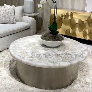 Op de afbeelding: Een witte agaat salontafel met een rond blad en een zilveren onderstel. De tafel staat op een wit, pluche vloerkleed. Een witte orchidee plant staat in een witte kom op de tafel.