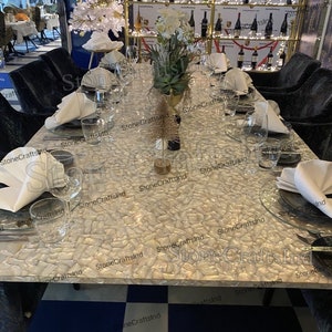 May include: A white table with a white mother of pearl mosaic tabletop. The table is set with white napkins, clear glasses, and silver plates. The table is surrounded by black chairs. The table is in a restaurant setting with a bar in the background.