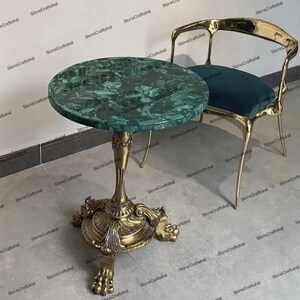 Op de afbeelding: Een ronde groene marmeren tafel met een gouden basis en ingewikkelde details. De tafel heeft drie poten met sierlijke voeten. De tafel staat op een lichtgrijze vloer.