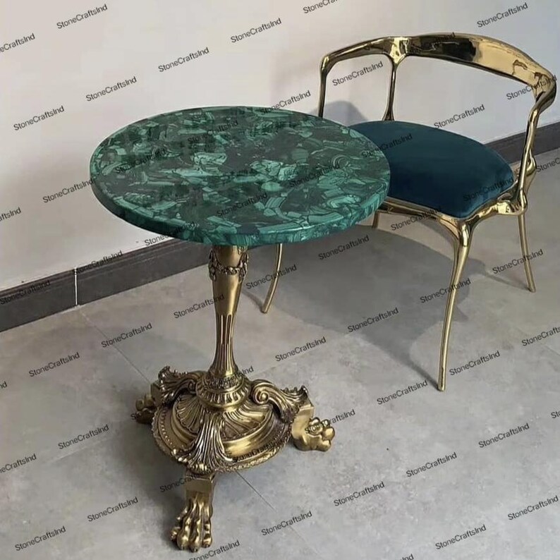 Op de afbeelding: Een ronde groene marmeren tafel met een gouden basis en ingewikkelde details. De tafel heeft drie poten met sierlijke voeten. De tafel staat op een lichtgrijze vloer.