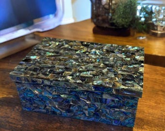 May include: A rectangular box with a lid, covered in iridescent mosaic tiles. The tiles reflect shades of blue, green, and black, creating a shimmering effect. The box is placed on a wooden surface.