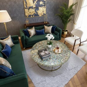 May include: A living room scene featuring a green velvet sofa and loveseat with blue and gold accent pillows. A round coffee table with a mosaic top sits on a gray rug. A gold floor lamp and a painting are also visible.