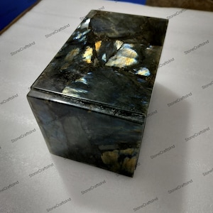 Op de afbeelding: Een rechthoekige Labradoriet doos met een gepolijste afwerking. De steen heeft een donkere basiskleur met iriserende flitsen van blauw, goud en groen. De doos heeft een solide, geometrische vorm en toont de natuurlijke schoonheid van de steen.